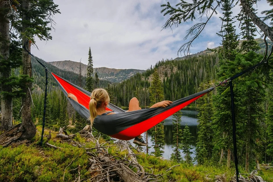Woman in red and black hammock between two trees looking at lake and trees